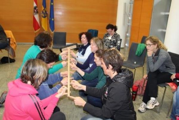 Eine Gruppe erwachsener Damen hält gemeinsam einen langen Holzstock in Händen