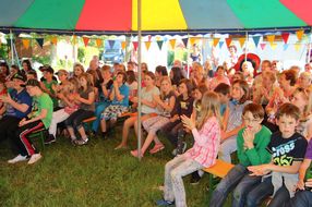 Viele Kinder sitzen auf den Bänken im bunten Zelt und hören begeistert zu.