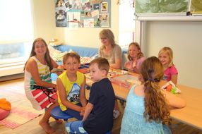 Sechs Kinder und eine Betreuerin sitzen um einen Tisch herum und spielen Monopoly.